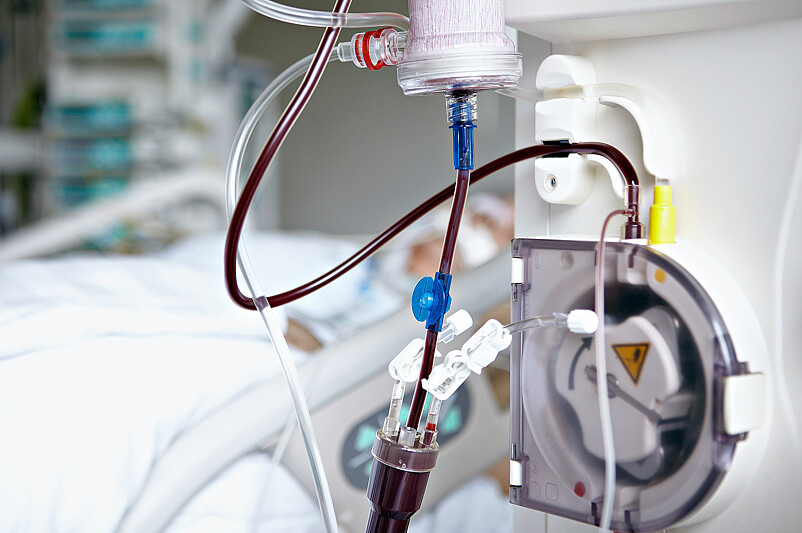 Es ist ein Gerät zur Apherese zu sehen, einem Verfahren, in dem Stammzellen aus dem Blut eines Spenders entnommen werden. Im Hintergrund ist verschwommen ein Patient im Krankenbett zu sehen.