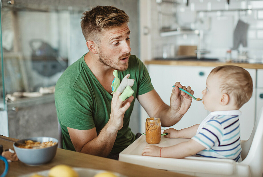 Ein junger Mann füttert ein Baby, das in einem Hochstuhl sitzt, mit Brei.