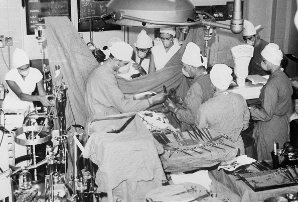 Die schwarz-weiß Aufnahme zeigt den Chirurgen Christiaan Barnard und sein OP-Team während einer Operation im Operationssaal. Alle haben ihren Blick auf den Patienten gerichtet und tragen OP-Kleidung samt weißer Hauben. Der Patient ist abgedeckt. Medizinische Geräte und OP-Besteck sind zu sehen.