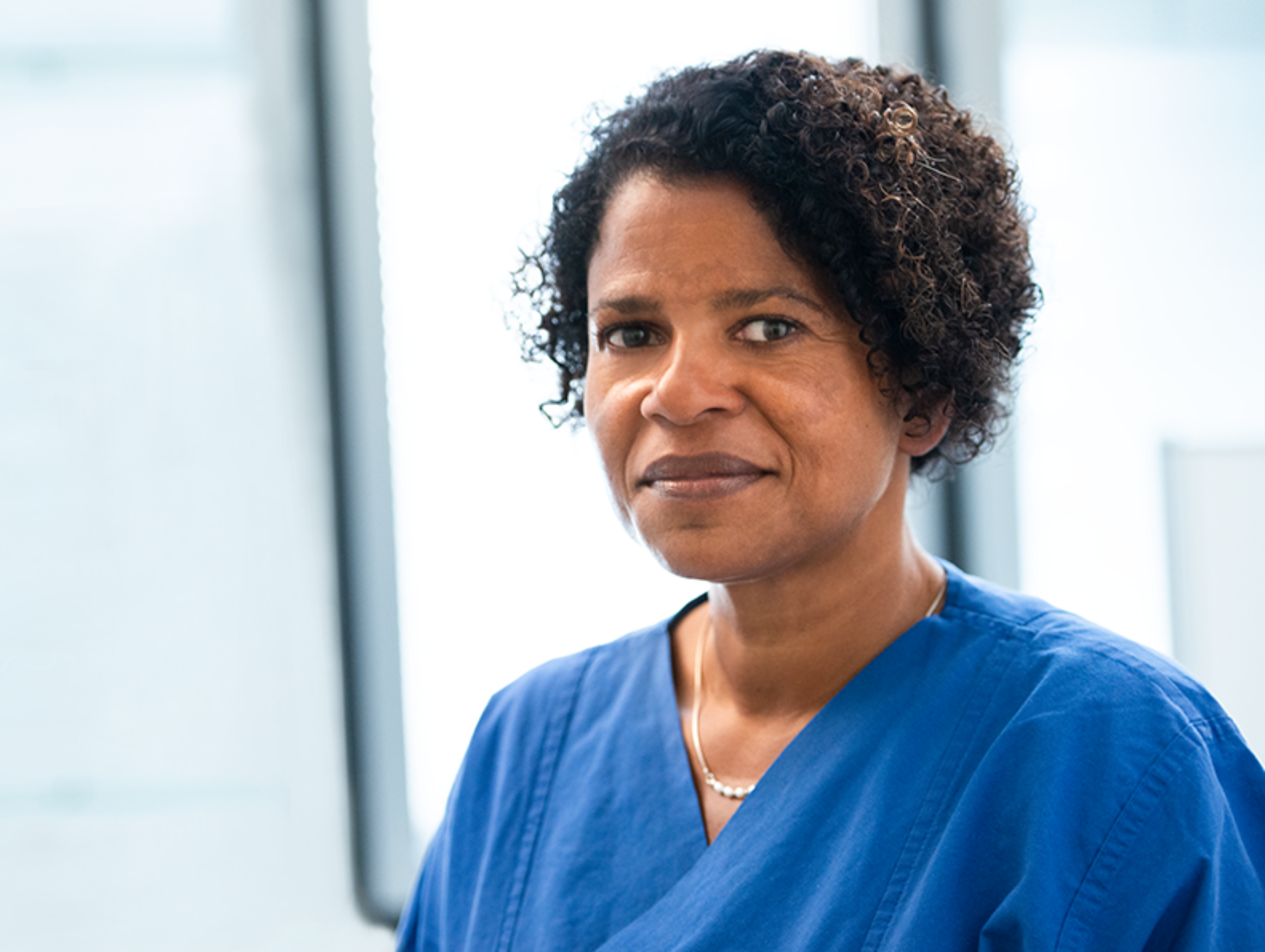 Das Foto ist eine Porträtaufnahme von Dr. Geraldine de Heer. Sie trägt medizinische Kleidung und lächelt in die Kamera.