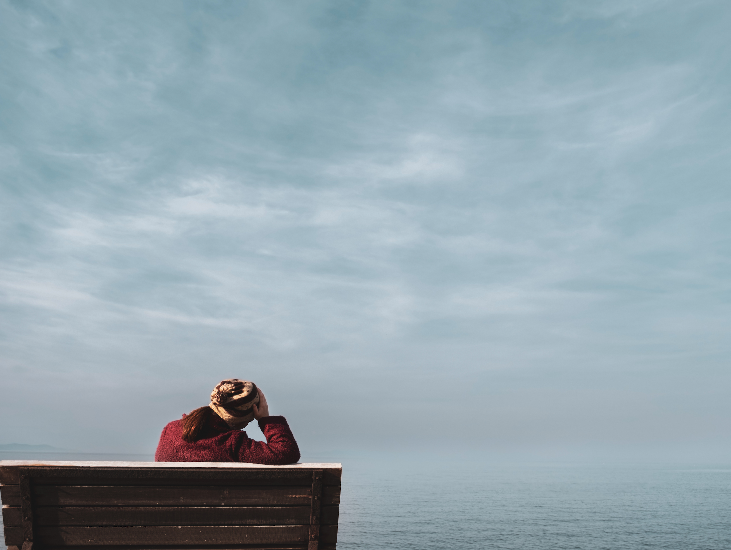 Eine Frau sitzt auf einer Bank. Sie hat ihren rechten Arm auf der Rücklehne abgestützt und hält ihren Kopf. Sie ist von hinten zu sehen. Im Hintergrund sieht man das Meer und den Himmel.