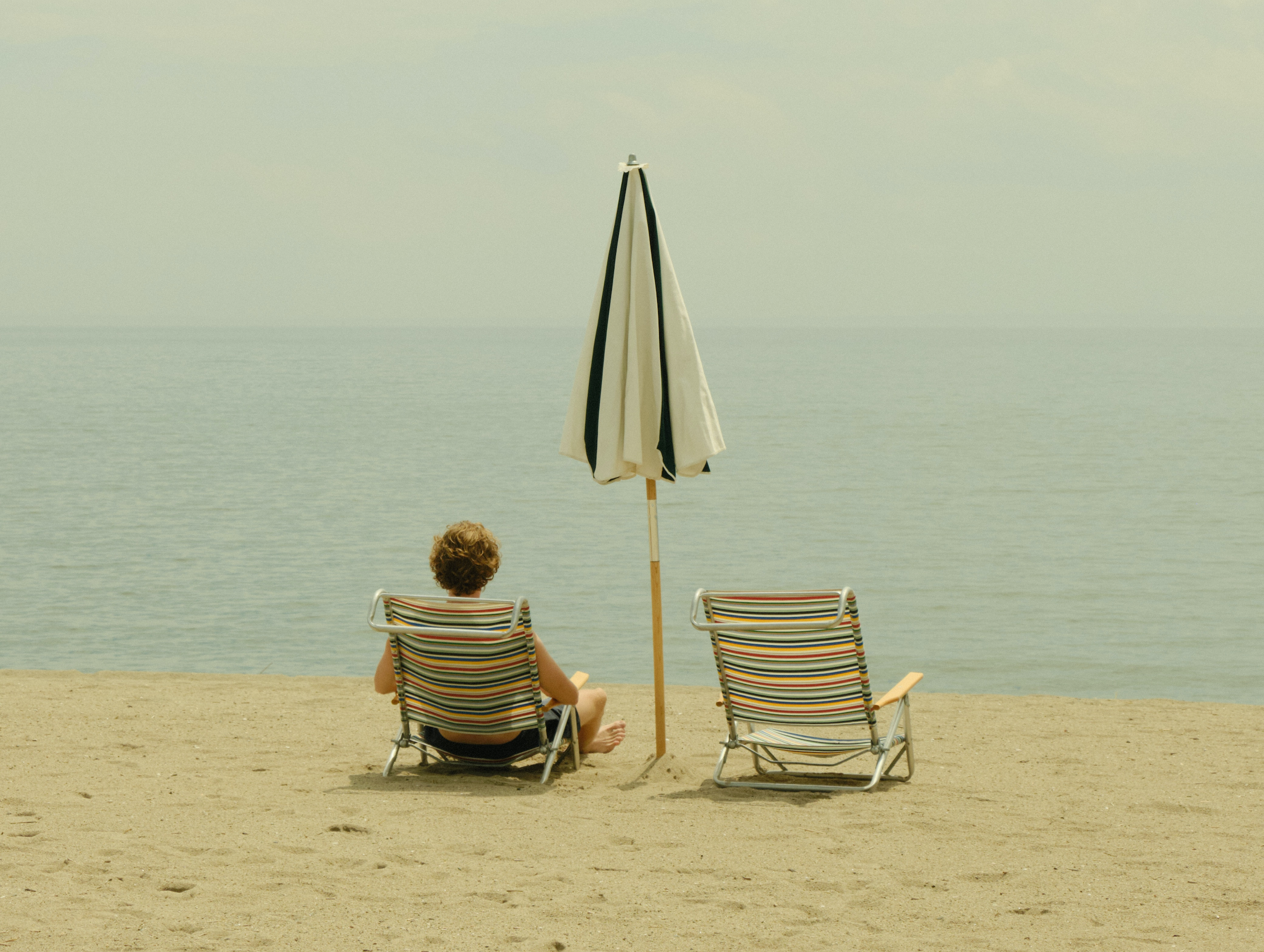 Eine Person liegt am Strand auf einer Liege. Eine weitere Liege rechts neben ihr ist frei. Dazwischen steht ein Sonnenschirm, der nicht aufgeklappt ist. Die Person schaut aufs Meer.