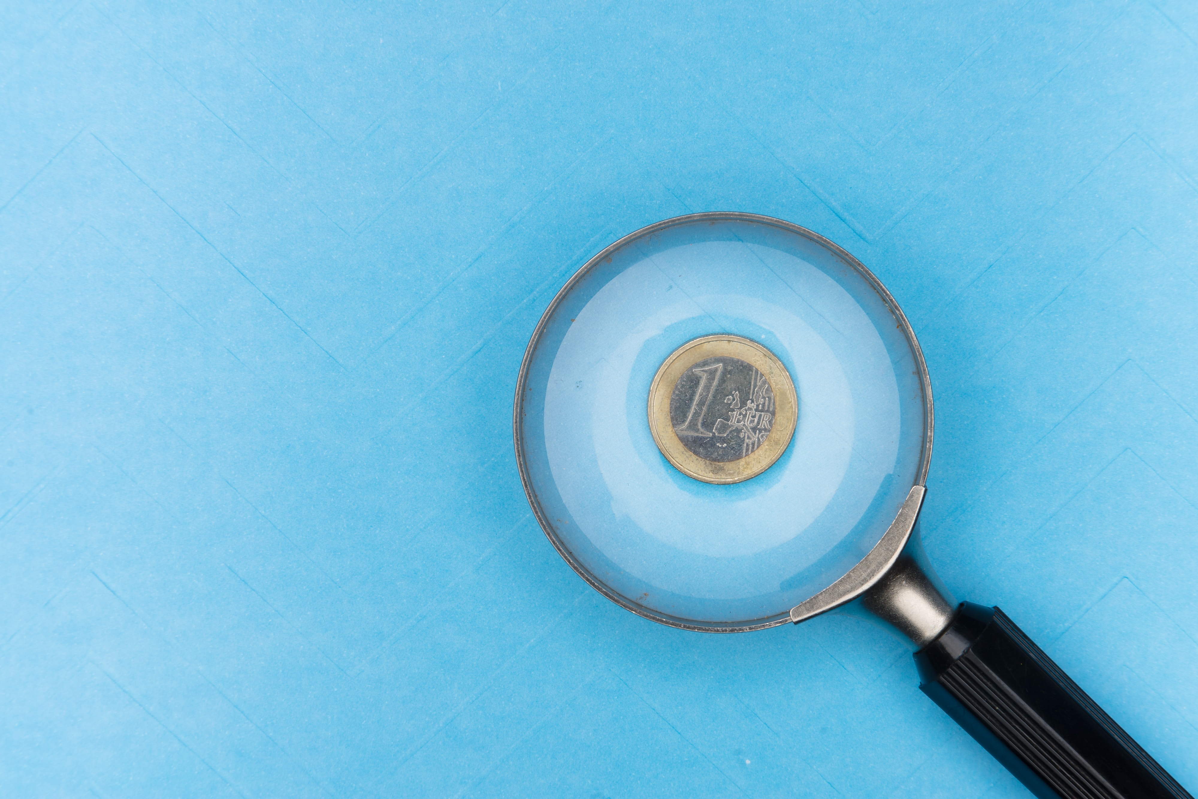 Eine Lupe liegt auf einer Ein-Euro-Münze auf blauem Hintergrund.