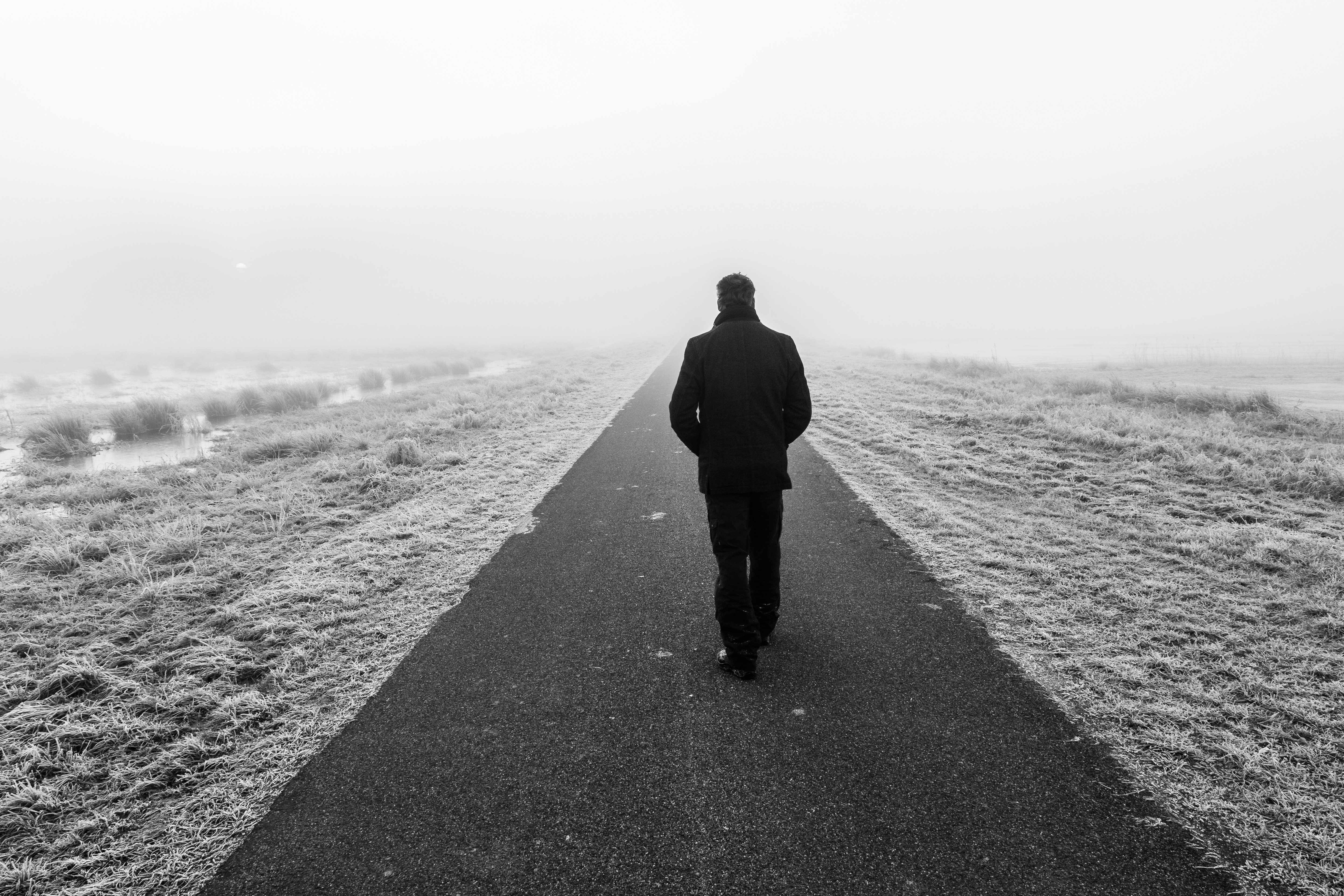 Das schwarz-weiß Foto zeigt einen Mann, der einen geteerten Weg zwischen zwei Feldern entlangläuft. Er läuft von der Kamera weg und ist mittig des Bildes. Seine Hände scheint er in den Jackentaschen zu haben.