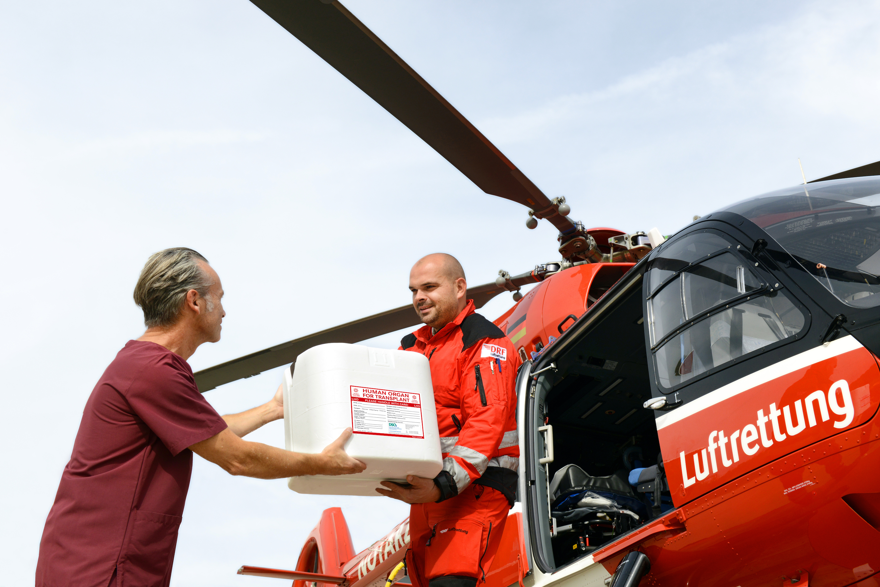 Ein Arzt gibt einem Sanitäter der Luftrettung eine Organ-Transportbox. Dahinter ist ein Hubschrauber der Luftrettung zu sehen.