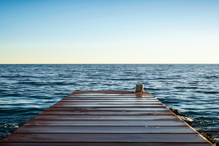 Auf dem Foto ist ein Holzsteg zu sehen, der ins Meer führt. Der Himmel ist blau.