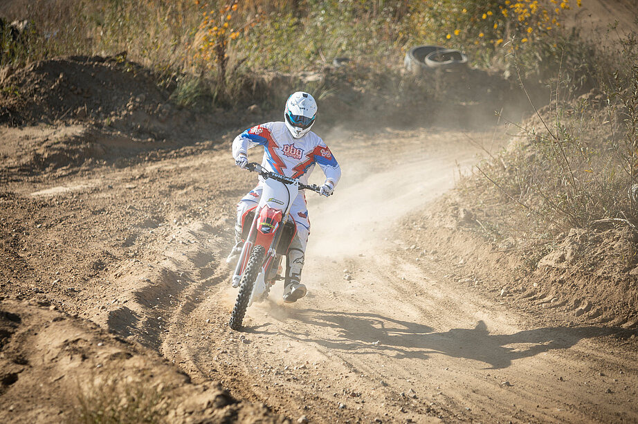 Herr Koch f&auml;hrt mit seinem Gel&auml;ndemotorrad einen sandigen Feldweg entlang. Er tr&auml;gt Motorradkleidung und eine Helm. Bei der Fahrt wirbelt er Staub auf.