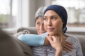 Zwei Frauen sitzen auf einem Sofa. Die Frau hinten legt den Arm um die Frau vor ihr. Beide schauen besorgt, aber auch hoffnungsvoll aus dem Fenster.