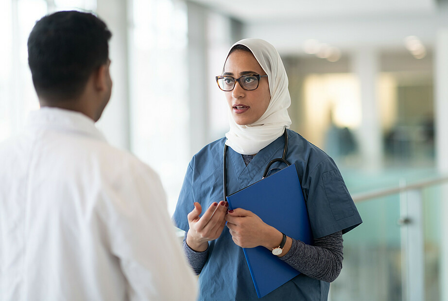 Zu sehen sind eine Ärztin, die ein Kopftuch trägt, und ein Arzt im Krankenhausflur, die im Gespräch sind. 