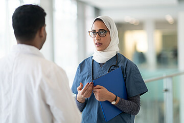 Zu sehen sind eine Ärztin, die ein Kopftuch trägt, und ein Arzt im Krankenhausflur, die im Gespräch sind. 