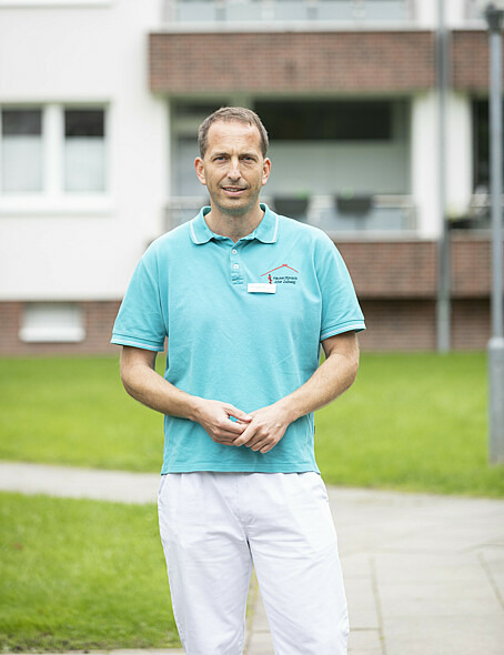 Dr. Michael von Bezold steht draußen und schaut in die Kamera. Hinter ihm sind eine Grünfläche und eine Hausfassade zu erkennen. Er trägt Arztkleidung und hält seine Hände vor seinem Bauch zusammen.