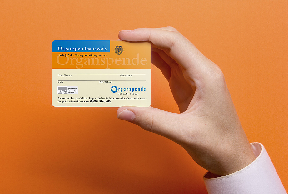 Ein Organspendeausweis wird von einer Hand hoch und frontal in die Kamera gehalten. Der Hintergrund ist orange.