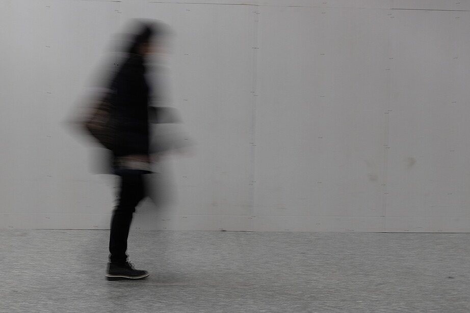 Eine schwarz gekleidete Person geht vor einer kahlen Betonwand entlang. Sie ist nur verschwommen zu erkennen.