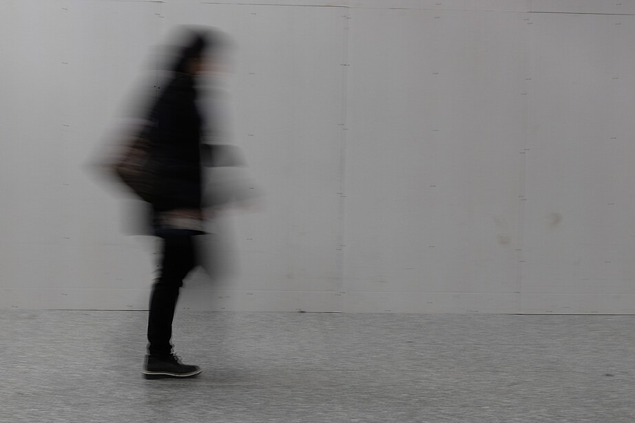 Eine schwarz gekleidete Person geht vor einer kahlen Betonwand entlang. Sie ist nur verschwommen zu erkennen.