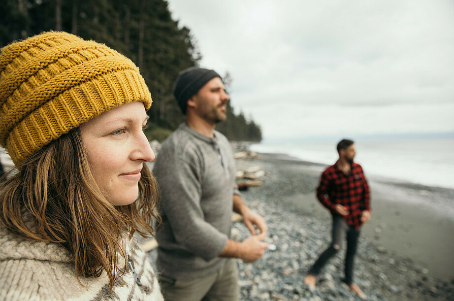 Drei Menschen stehen am Ufer eines großen Gewässers. Im Vordergrund ist eine lächelnde Frau zu sehen, die eine knallgelbe Mütze trägt. Im Hintergrund schnippsen zwei Männer Steine ins Wasser.