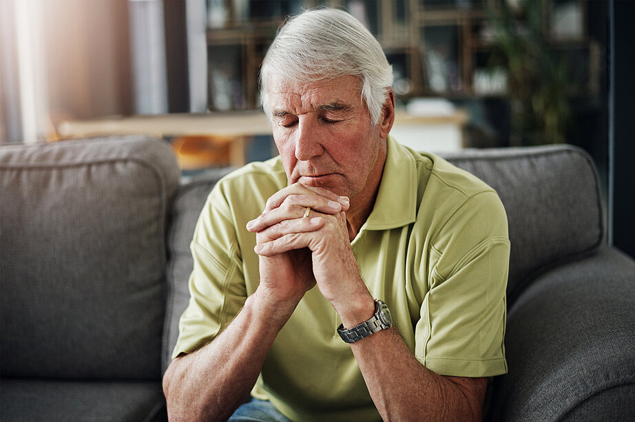 Zu sehen ist ein älterer Mann, der seine Hände ineinander gefaltet hat und im Gebet vertieft ist. Dabei hat er seine Augen geschlossen. Er sitzt auf einem Sofa und hat seine Ellbogen auf die Knie gestützt.