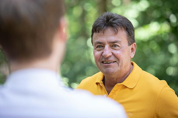 Herr Röschert ist im Gespräch. Man sieht ihn von vorn, er lächelt. Im Vordergrund steht eine weitere Person, die von hinten zu sehen ist. 