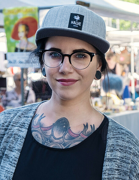 Die 33-jährige Nicci blickt grinsend in die Kamera. Sie trägt eine Brille, eine Kappe und ein dunkles Shirt, worunter Tattoos zu erkennen sind.