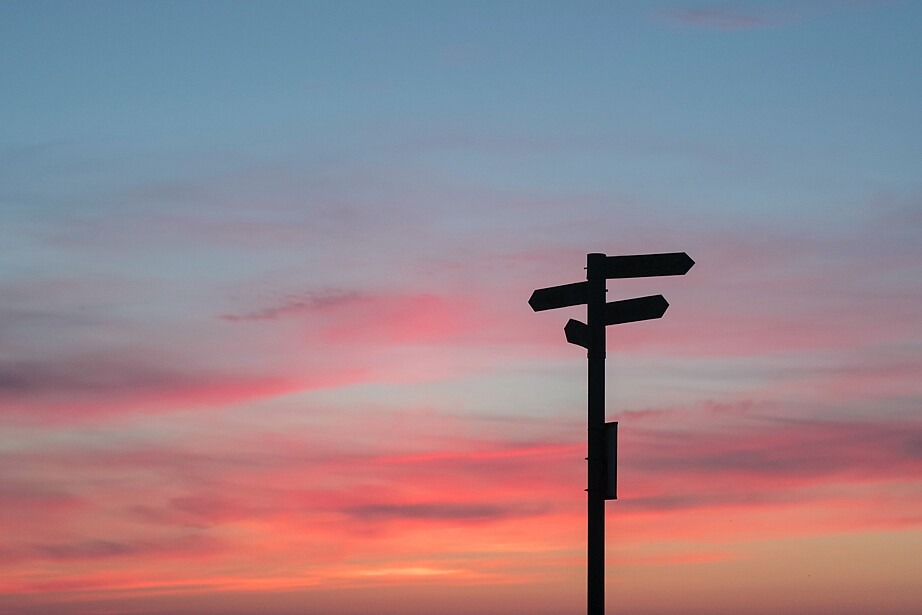 Ein Straßenschild mit mehreren Abzweigungen in unterschiedliche Richtungen ist vor einem rosa gefärbtem Himmel zu sehen.