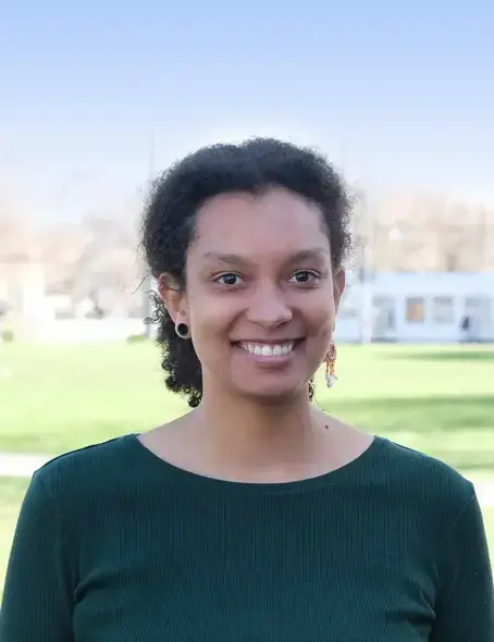 Die 22-j&auml;hrige Alissa steht drau&szlig;en vor einer Wiese und blickt l&auml;chelnd nach vorne. Sie hat ihre dunklen Locken zu einem Zopf zusammengebunden und tr&auml;gt ein dunkelgr&uuml;nes Shirt.