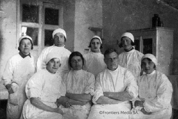 Es handelt sich um eine historische schwarz-weiß Aufnahme von dem ukrainischen Arzt Yu Yu Vorony und seinem OP-Team. Insgesamt sind acht Personen in weißer OP-Kleidung zu sehen, vier sitzen vorne, vier stehen dahinter. Alle schauen in die Kamera.