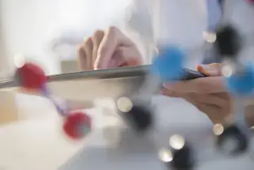 Die Hand eines Arztes ist im Hintergrund zu erkennen, wie sie auf ein Tablet tippt. Davor ist verschwommen ein Molek&uuml;lmodell zu sehen. 