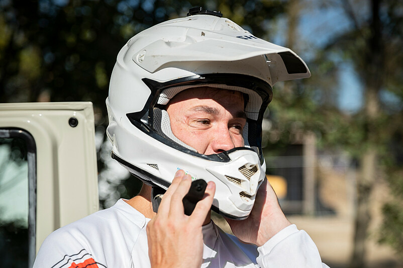 Herr Koch trägt Motorradkleidung. Er hält die Hände an seinen Motorcross-Helm, als wolle er ihn auf- oder absetzen.