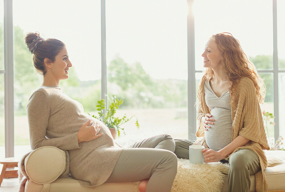 Zwei schwangere Frauen sitzen auf einer gepolsterten Bank vor einem Fenster und unterhalten sich. Die Sonne scheint, beide lächeln und sehen entspannt aus.