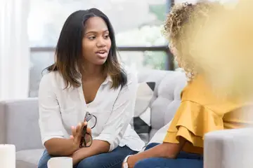 Zwei Frauen sitzen auf einem Sofa und unterhalten sich angeregt. Eine der Frauen ist von vorne zu sehen, sie h&auml;lt eine Brille in der Hand und gestikuliert. Die andere Frau ist von der Seite zu sehen.