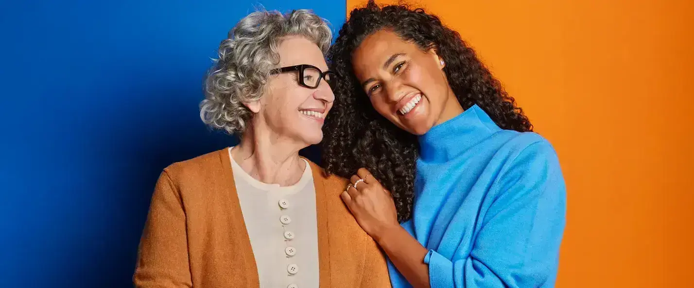 Zu sehen sind eine &auml;ltere Frau mit orangenem Cardigan auf blauem Hintergrund und eine j&uuml;ngere Frau mit einem blauen Pullover auf orangenem Hintergrund. Beide Frauen l&auml;cheln und stehen vertraut aneinander.