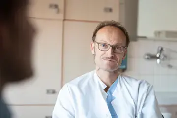 Dr. Frank Logemann im Gespr&auml;ch mit einem Patienten, der im Vordergrund nur als Umriss erkennbar ist. Herr Dr. Logemann h&ouml;rt aufmerksam zu.