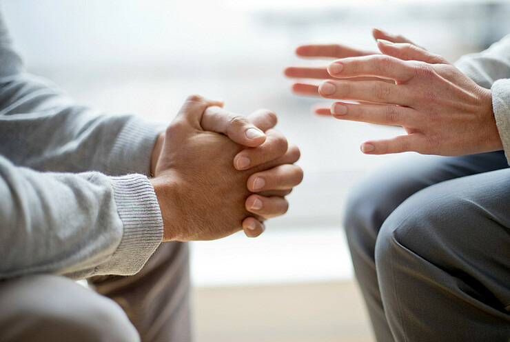 Zwei Personen sitzen sich gegenüber. Der Bildausschnitt zeigt nur ihre Knie und Unterarme. Die linke Person hat ihre Unterarme aufgestützt und die Hände gefaltet. Die rechte Person hält ihre Hände dagegen offen, als würde sie gestikulieren.
