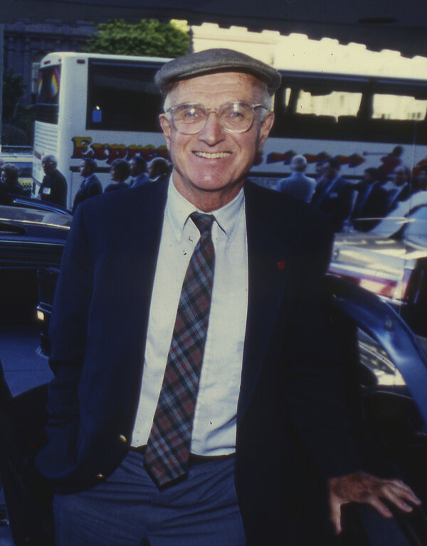 Das Foto zeigt den US-amerikanischen Chirurgen Joseph E. Murray in älteren Jahren. Es scheint sich eher um eine Aufnahme im Freizeitkontext zu handeln. Er hat die Hände hinter seinem Rücken verschränkt und lächelt glücklich in die Kamera. Er trägt einen Anzug mit Krawatte, eine Brille und eine Schiebermütze. Im Hintergrund ist ein Tourismus-Bus mit einer kleinen Ansammlung an Menschen davor zu erkennen.