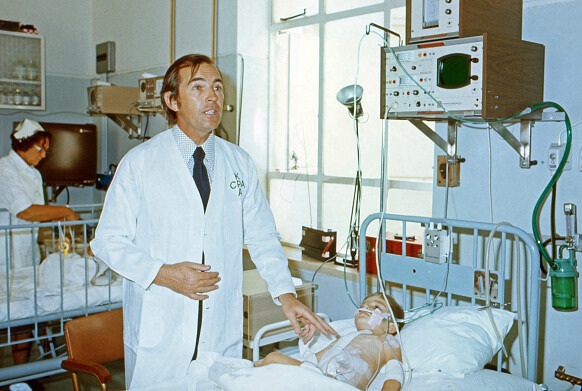 Das Farbfoto zeigt den Chirurgen Christiaan Barnard in weißem Kittel an einem Patientenbett in einem Krankenzimmer. Der Patient ist ein kleiner Junge, der an Schläuchen im Bett liegt. Christiaan Barnard redet und gestikuliert mit seinen Händen. Im Hintergrund ist eine Krankenpflegerin mit weißer Haube an einem weiteren Krankenbett zu erkennen.