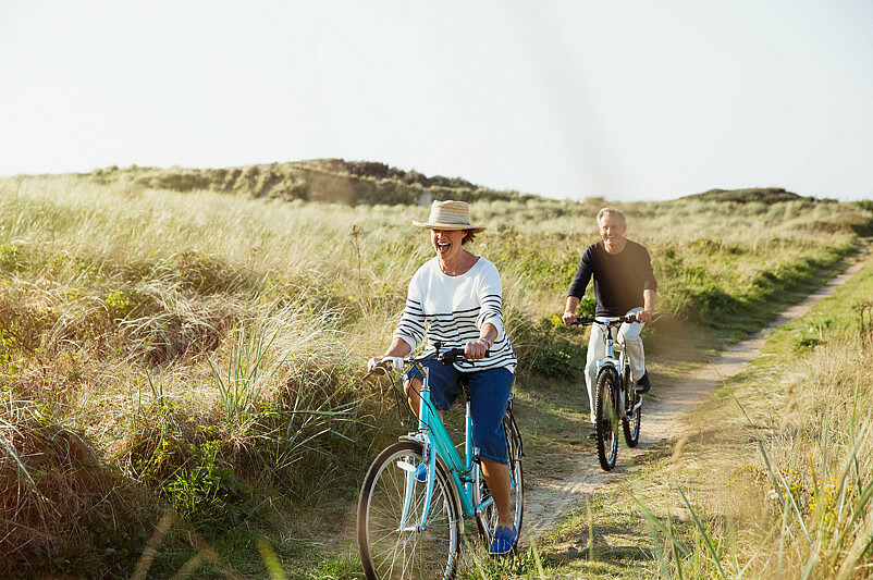 Eine Frau und ein Mann fahren auf Fahrrädern einen Feldweg entlang. Beide tragen Freizeitkleidung und lachen. Die Sonne scheint.