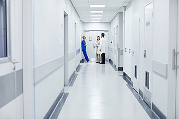 Am Ende eines Krankenhausflurs stehen eine Krankenhausmitarbeiterin in OP-Kleidung und eine Ärztin und ein Arzt in weißen Kitteln und beraten sich.