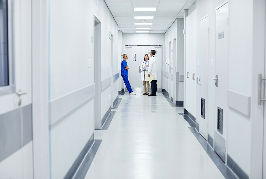 Am Ende eines Krankenhausflurs stehen eine Krankenhausmitarbeiterin in OP-Kleidung und eine Ärztin und ein Arzt in weißen Kitteln und beraten sich.