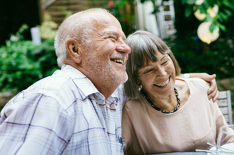 Ein Ehepaar im Seniorenalter sitzt gemeinsam an einem Tisch. Er hält sie im Arm und beide lachen herzlich und mit geschlossenen Augen.