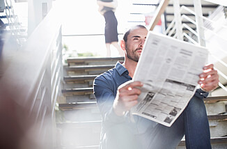 Zu sehen ist ein Mann, der auf den Stufen einer Treppe sitzt und aufmerksam eine Zeitung liest.