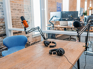 Zu sehen ist ein Podcaststudio. Auf einem Tisch liegen zwei Kopfhörer. Außerdem ist ein orangenes Mikro angebracht. Davor steht ein blauer Stuhl. Im Hintergrund ist hinter einer Glasscheibe ein Schreibtisch mit Bildschirmen zu erkennen.