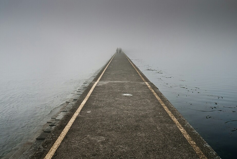 Ein langer geteerter Weg führt zum Horizont hin in den Nebel. Weit hinten sind Personen zu erkennen. Links und rechts ist Wasser.