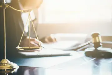 Auf dem Foto ist ein Richtertisch zu sehen. Im Hintergrund sitzt eine Richterin, vor ihr liegt der f&uuml;r den Gerichtsaal klassische Hammer. Im Vordergrund steht eine Waage.