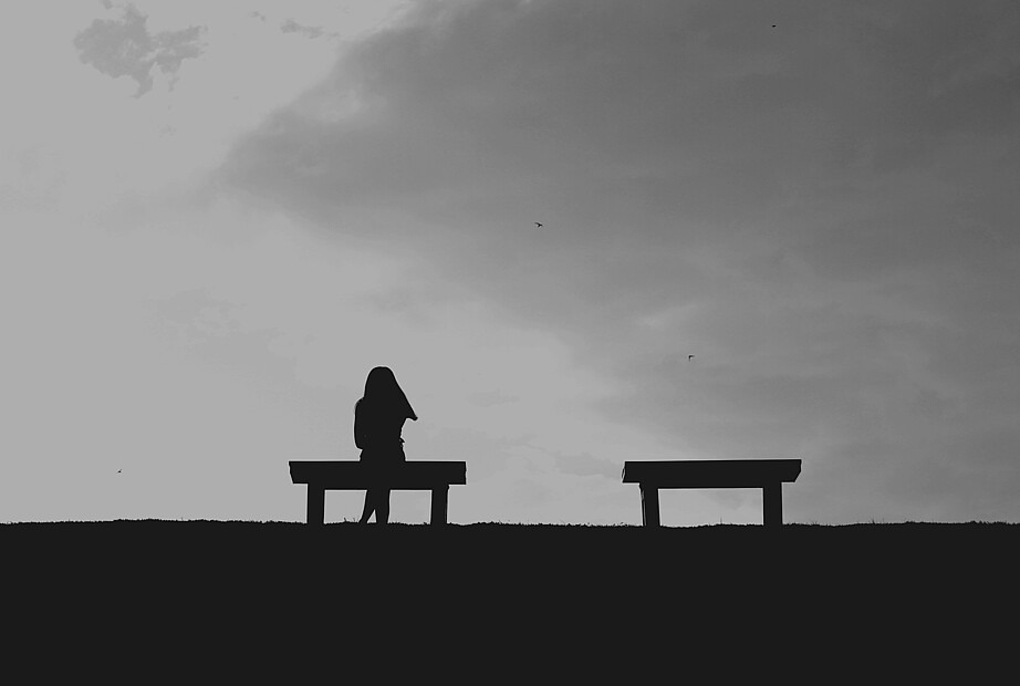 Auf dem schwarz-weiß Foto stehen zwei Bänke nebeneinander. Auf der linken sitzt eine Person. Es ist lediglich ihre Silhouette auszumachen.