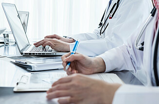 Zwei medizinische Fachkräfte sitzen an einem Tisch und arbeiten. Ein Mediziner tippt etwas in seinen Laptop ein, während die andere medizinische Fachkraft ein Formularblatt ausfüllt.