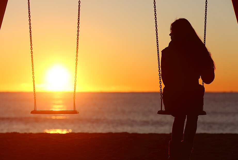 Eine Frau sitzt auf einer Schaukel. Sie schaut auf eine weitere Schaukel links von ihr, die leer ist. Im Hintergrund geht die Sonne über dem Meer unter.