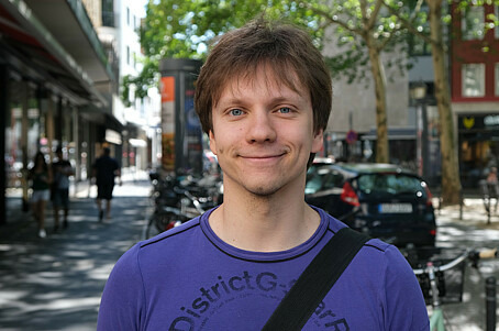 Der 26-jährige Dennis blickt in einer Straßenszene freundlich in die Kamera. Er hat dunkle kurze Haare und trägt ein violettes Shirt.