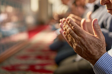 Zu sehen sind die Hände eines Mannes, die zum Gebet angehoben sind. Unscharf im Hintergrund sind der Innenraum einer Moschee und weitere kniende Menschen zu sehen. 
