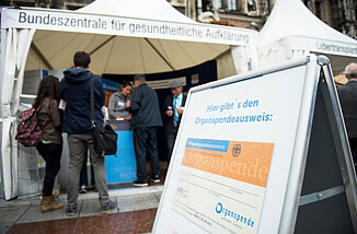 Zu sehen ist ein Klappschild, auf dem ein Organspendeausweis zu sehen ist. Es steht vor einem Informationsstand.