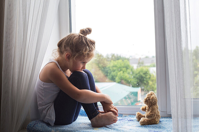 Das Bild zeigt ein trauriges Kind auf einem Fensterbrett und einen Teddybär.