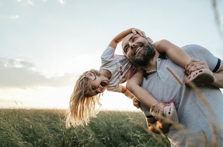 Ein Mann tr&auml;gt ein kleines M&auml;dchen auf den Schultern. Die Sonne scheint und beide lachen fr&ouml;hlich.